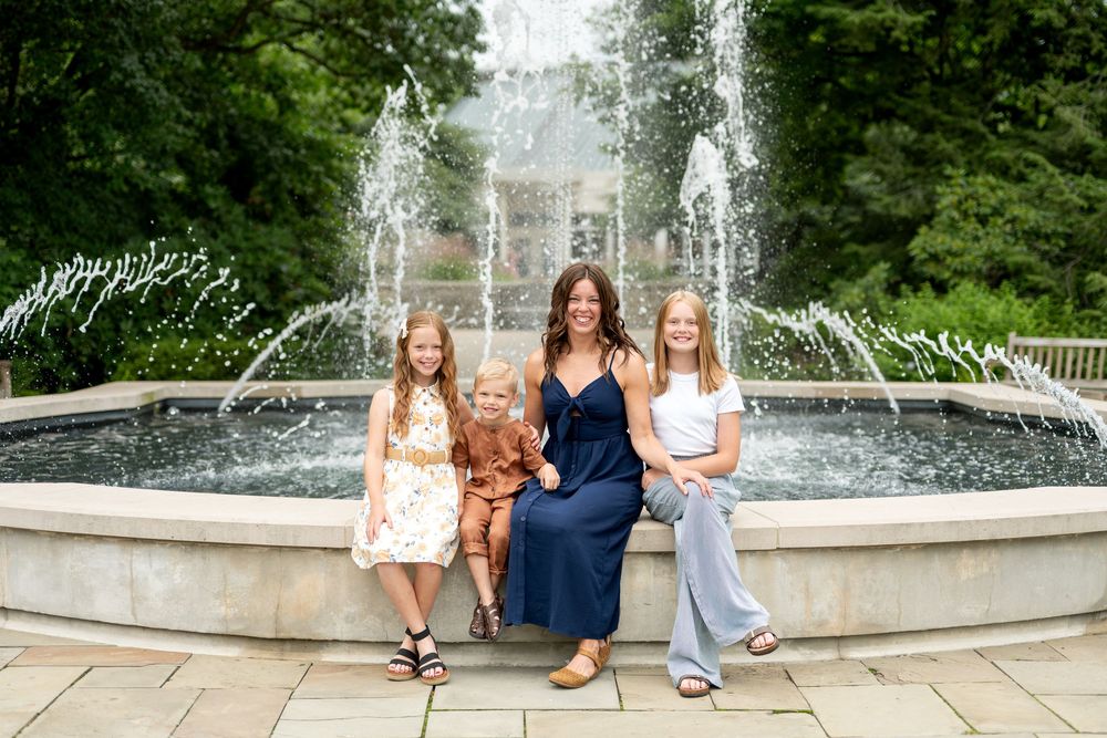 A woman and three girls smile in front of a fountain outdoors.