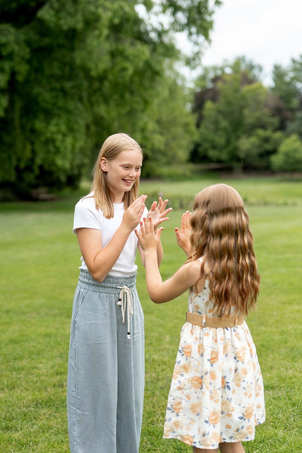 Two girls playing a clapping hand game together on green grass in a sunny park, wearing summer dresses.