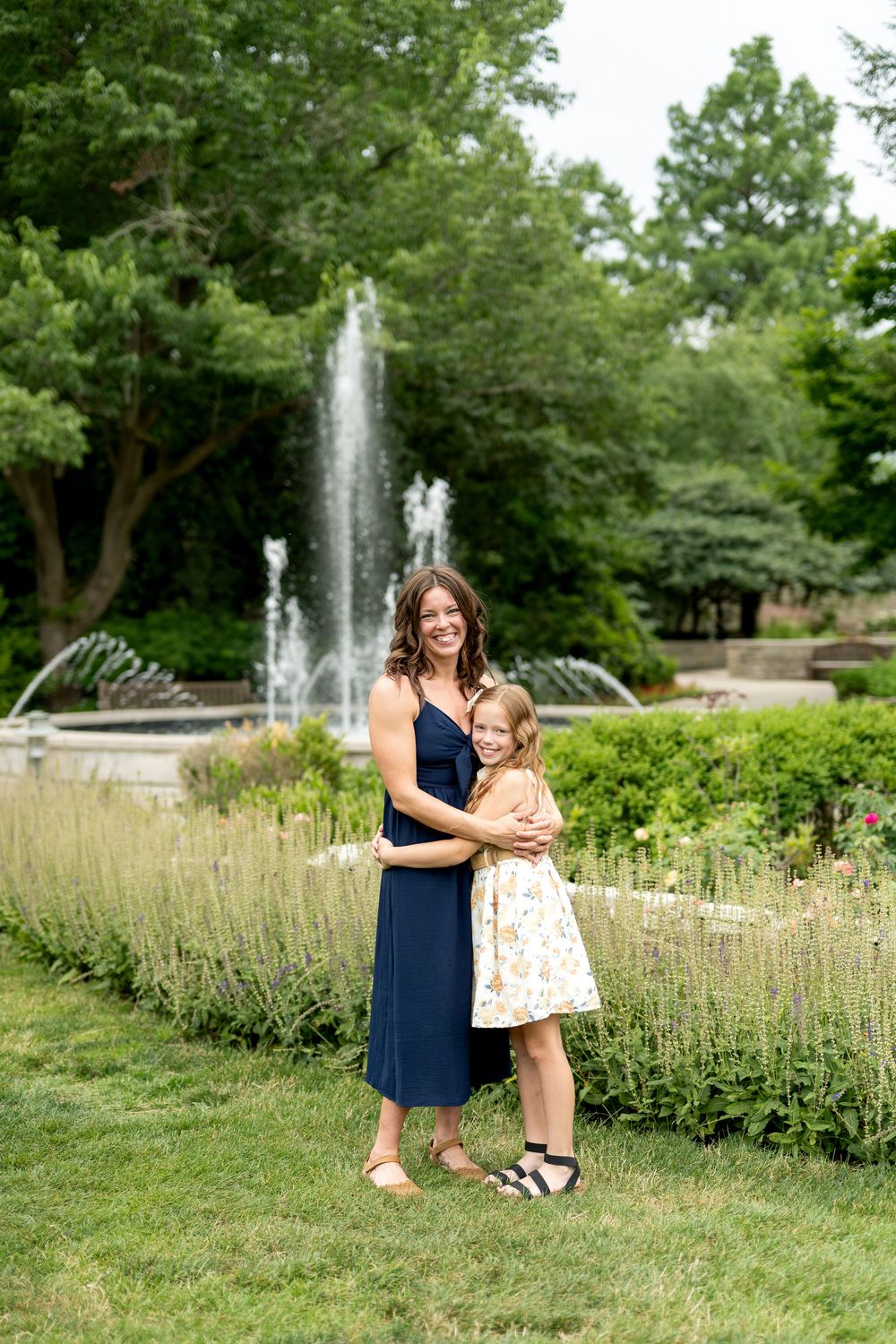 A mother in a navy dress embraces her young daughter in a floral dress near a park fountain.