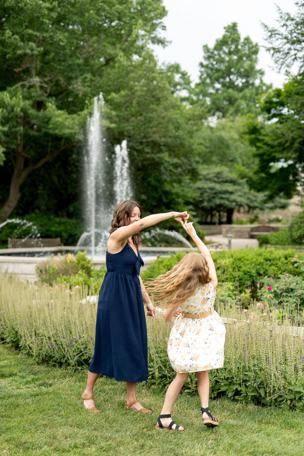 A woman in a navy dress twirls a girl in a floral dress in a lush garden park.