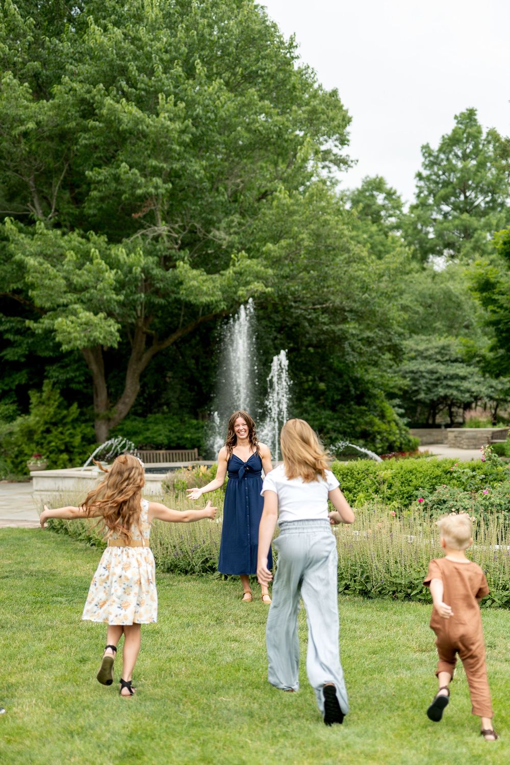 Three children run toward their smiling mother in a navy dress near a lush green park fountain.