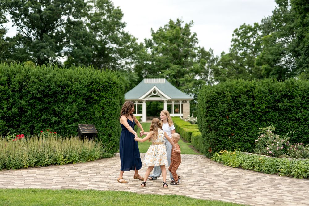 A mother in a navy dress walks with three children on a brick path toward a charming garden gazebo.