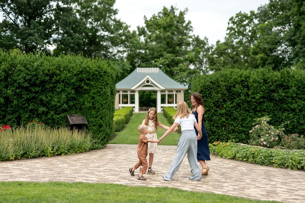 Two women and two children hold hands walking on a brick garden path with a green gazebo behind them.