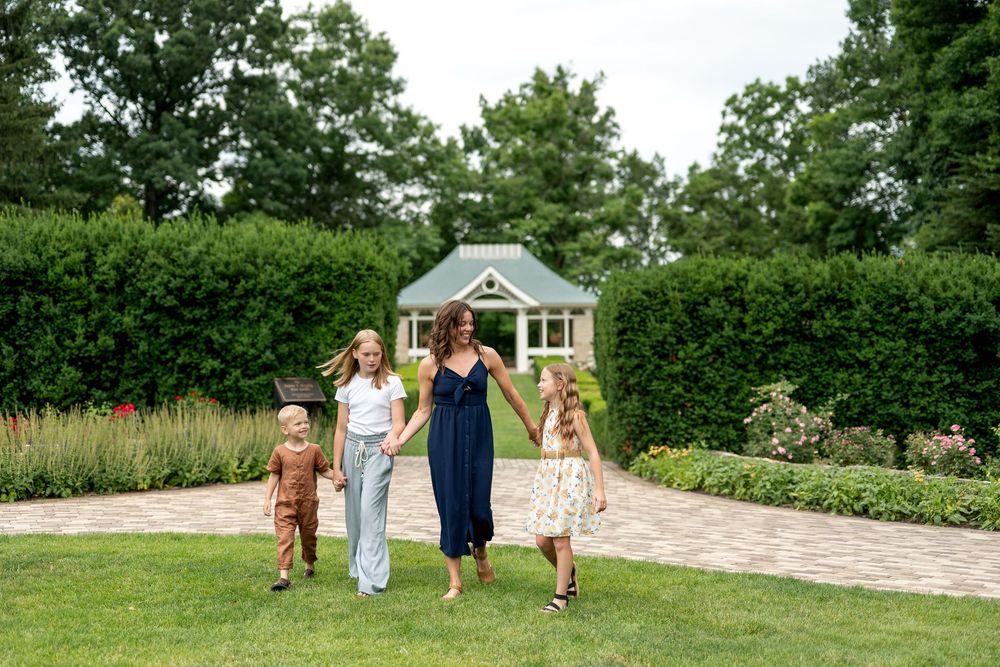 A mother in a navy dress walks hand-in-hand with three children on a brick path in a lush garden.