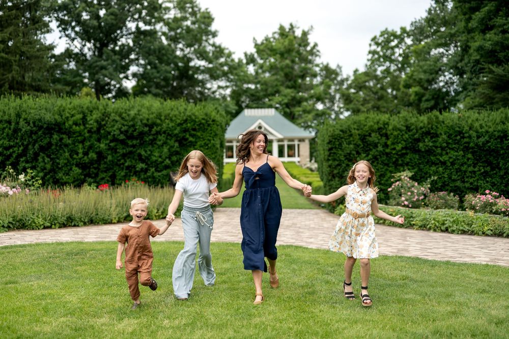 A mother and three children run joyfully across a green lawn in a garden with colorful flowers.