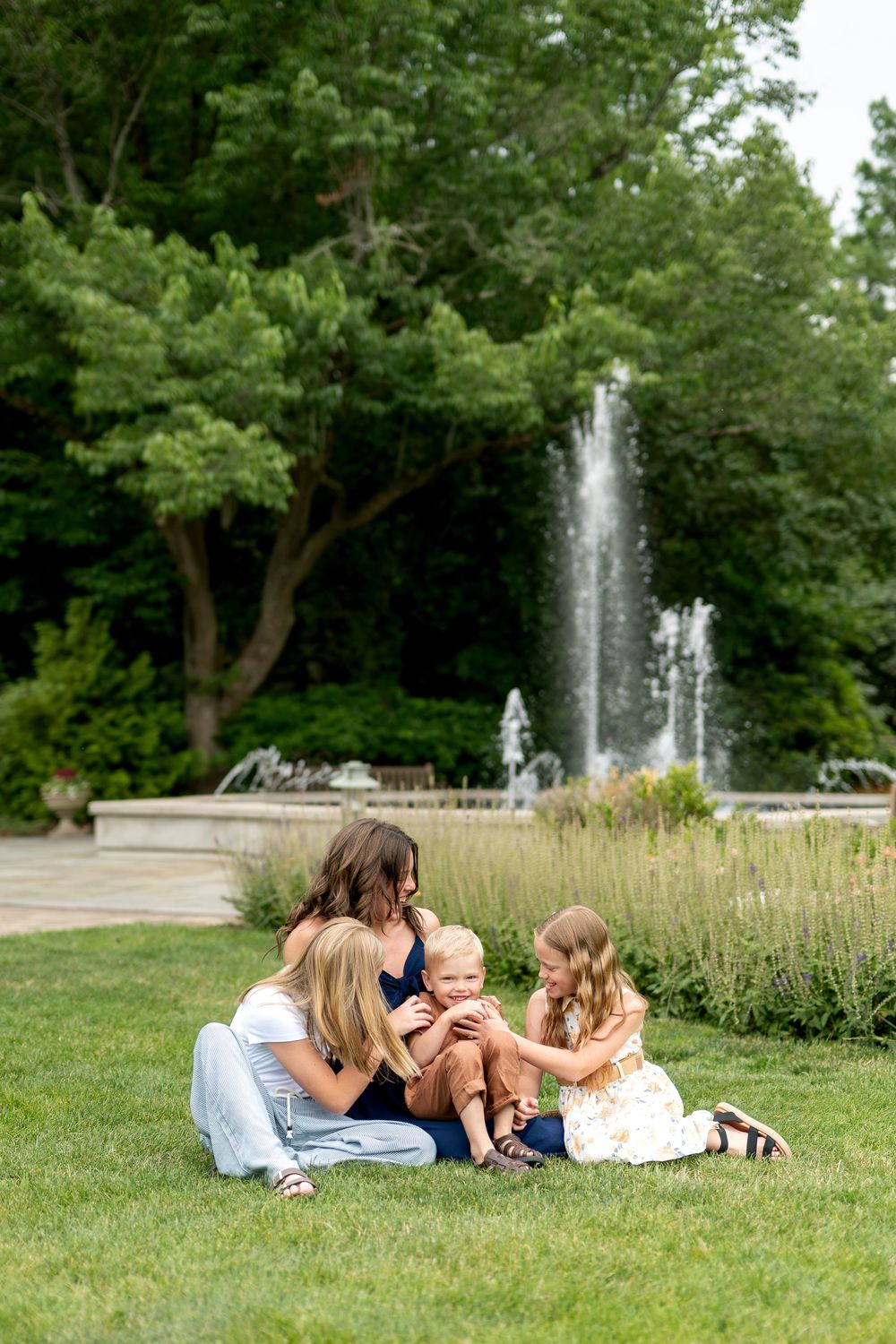 Family sits on grass near fountain as siblings interact with baby.