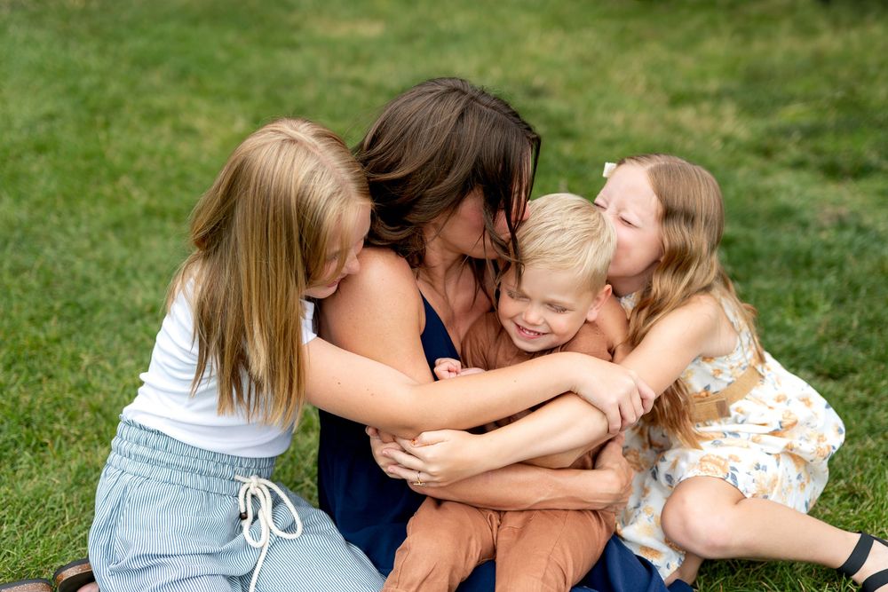 A mother sits on grass hugging three young children who lovingly embrace her outdoors.