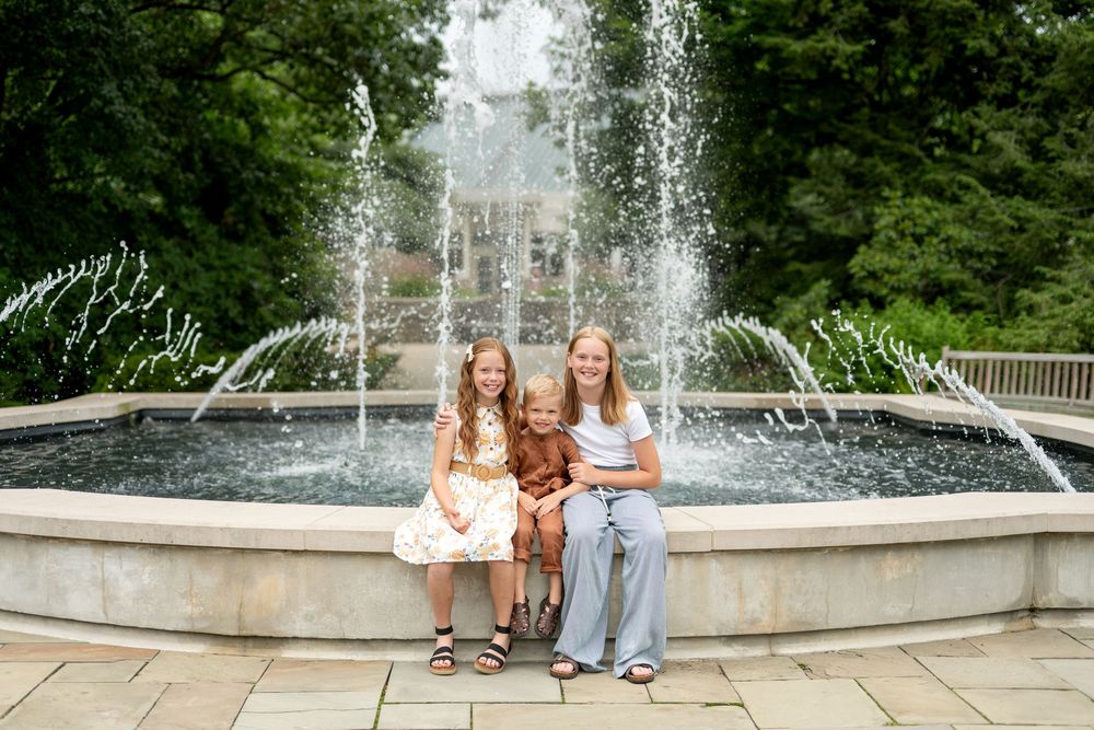 Two girls and a woman smile together in front of a large outdoor fountain surrounded by green trees.