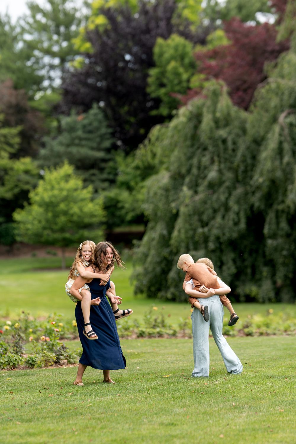 Two adults playfully carry laughing children in a lush green park setting.