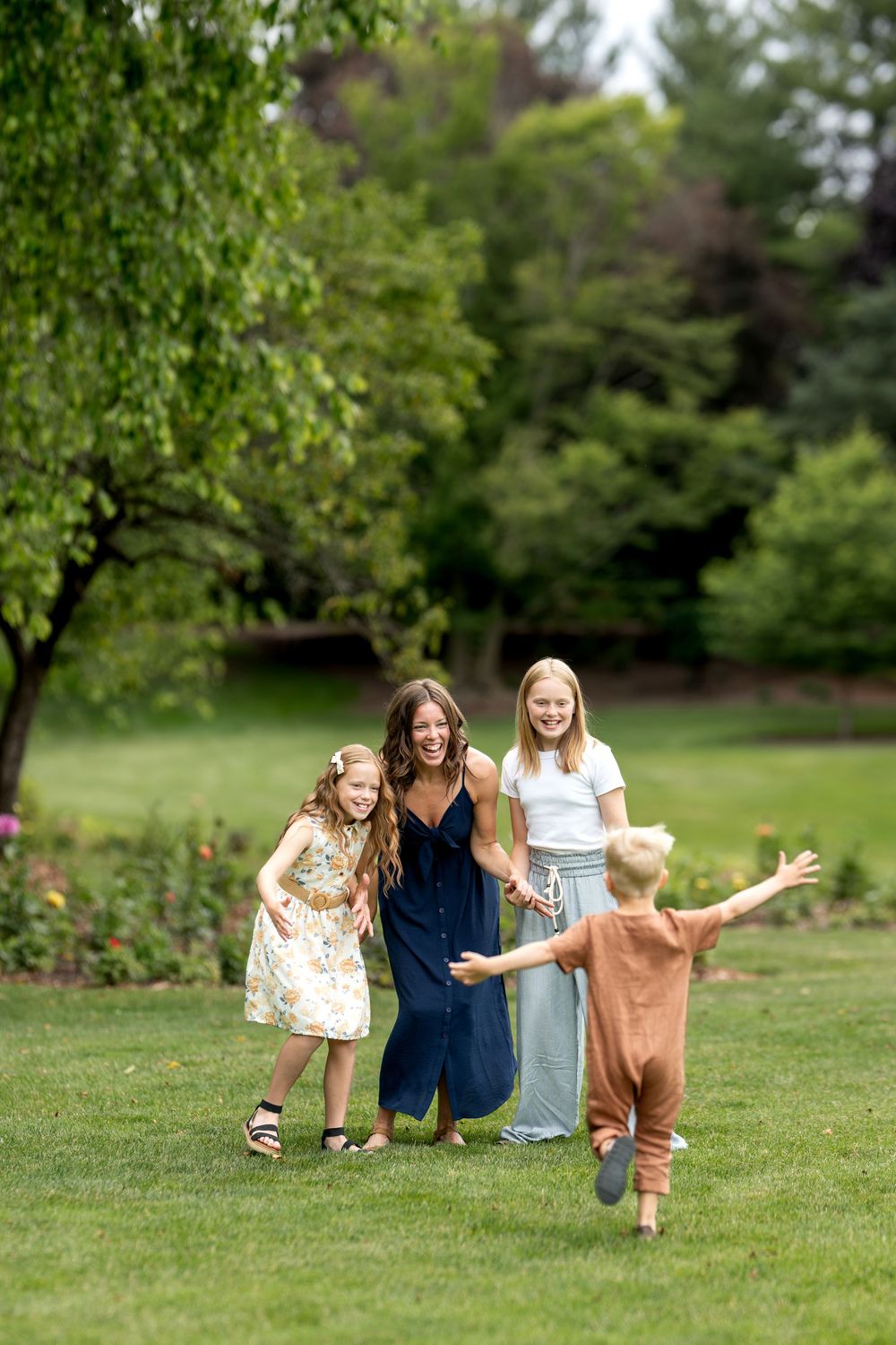 A laughing mother in navy stands with three children as a young boy runs toward them arms wide open.