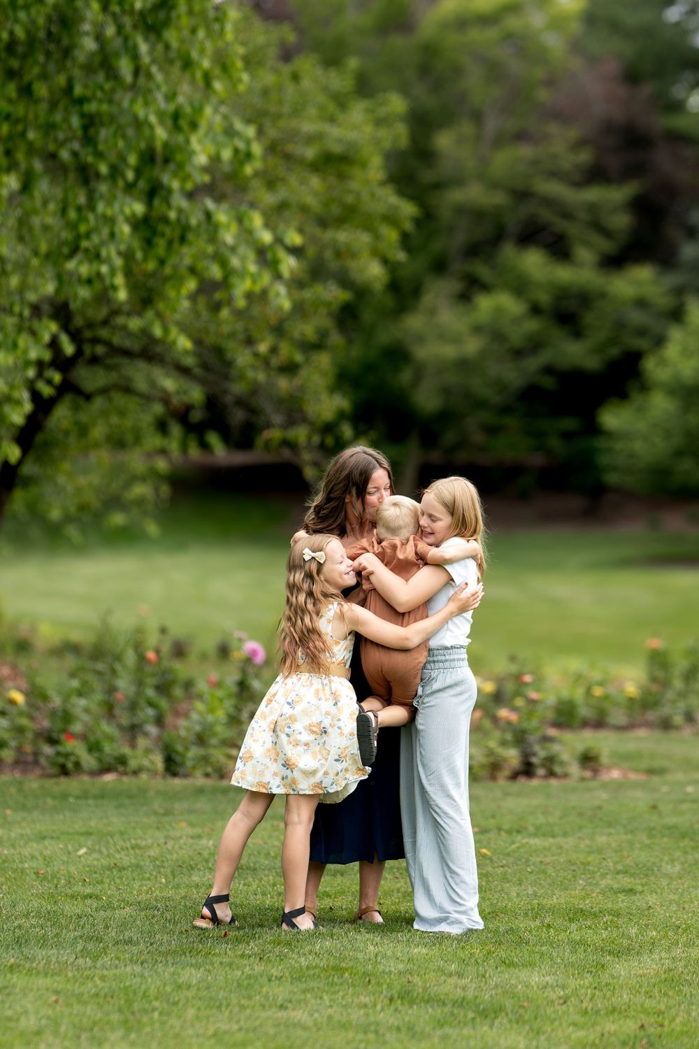 A mother hugs and kisses her young son while two daughters gather close in a lush green park.