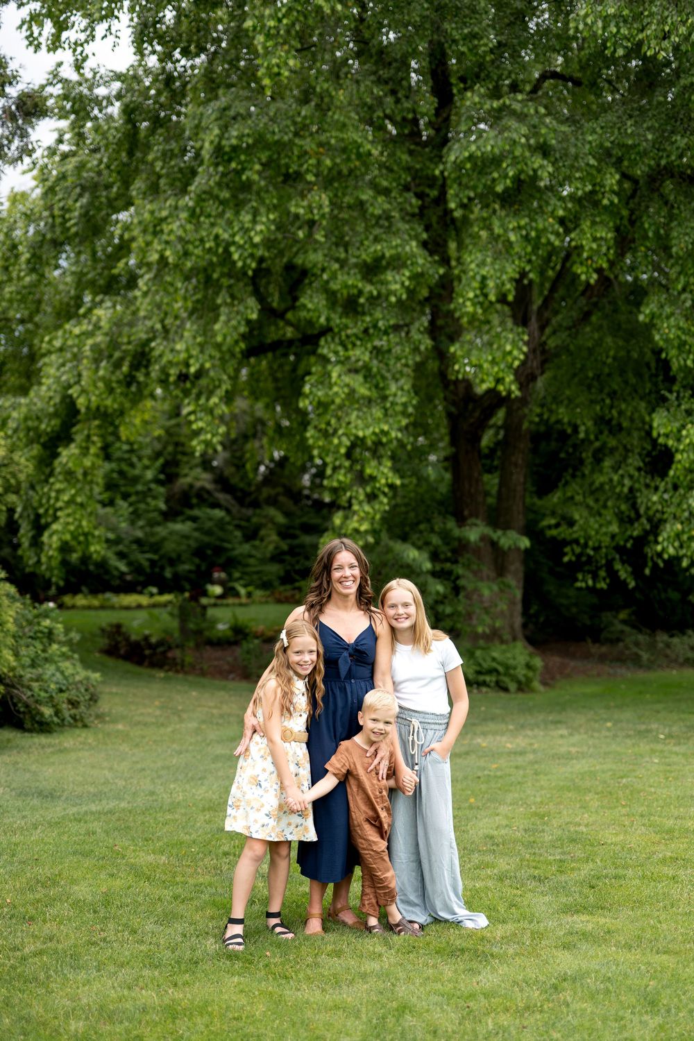 A smiling mother poses with her three children on a green lawn under a large tree.