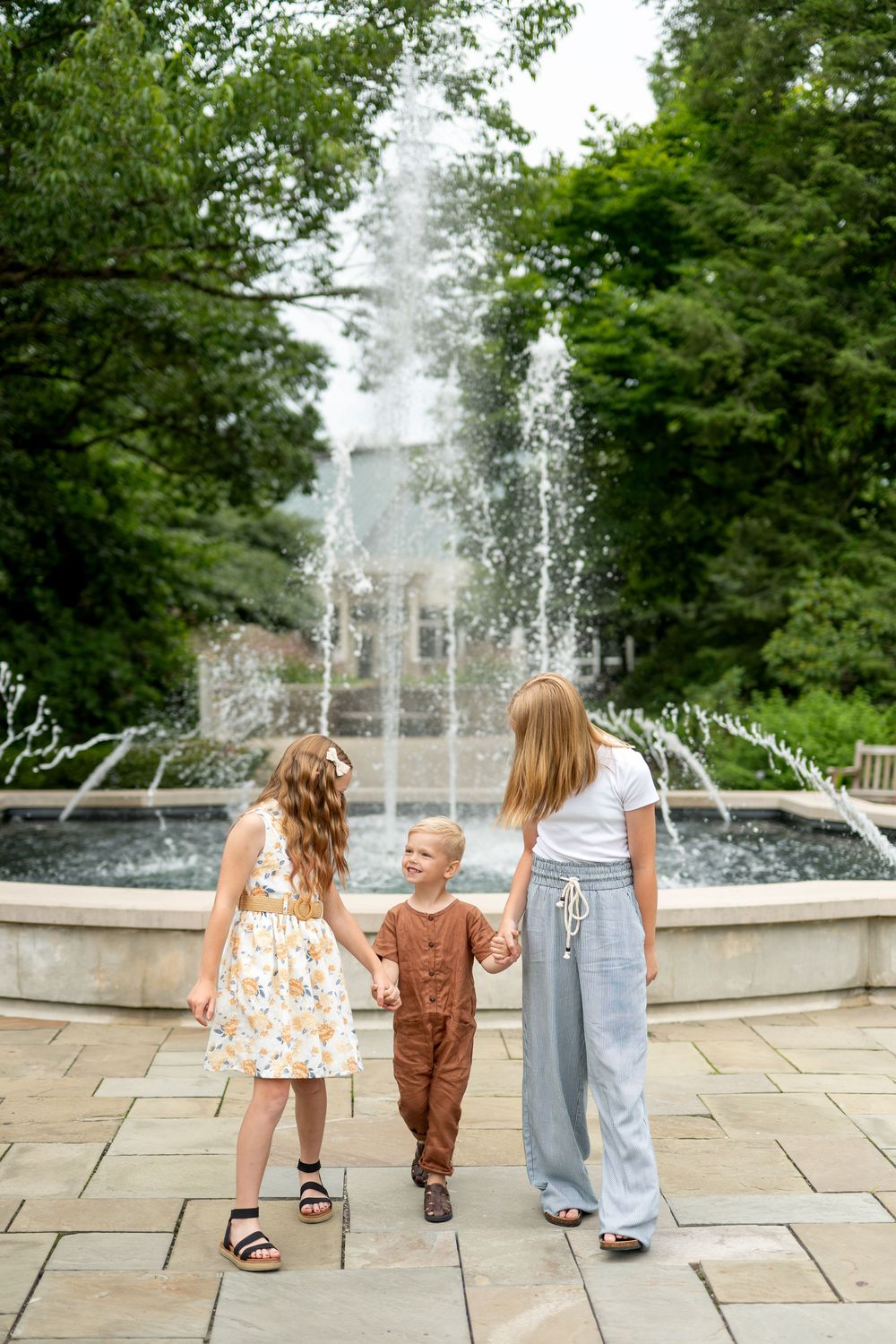 Three children hold hands in front of a large fountain surrounded by lush green trees in a park.