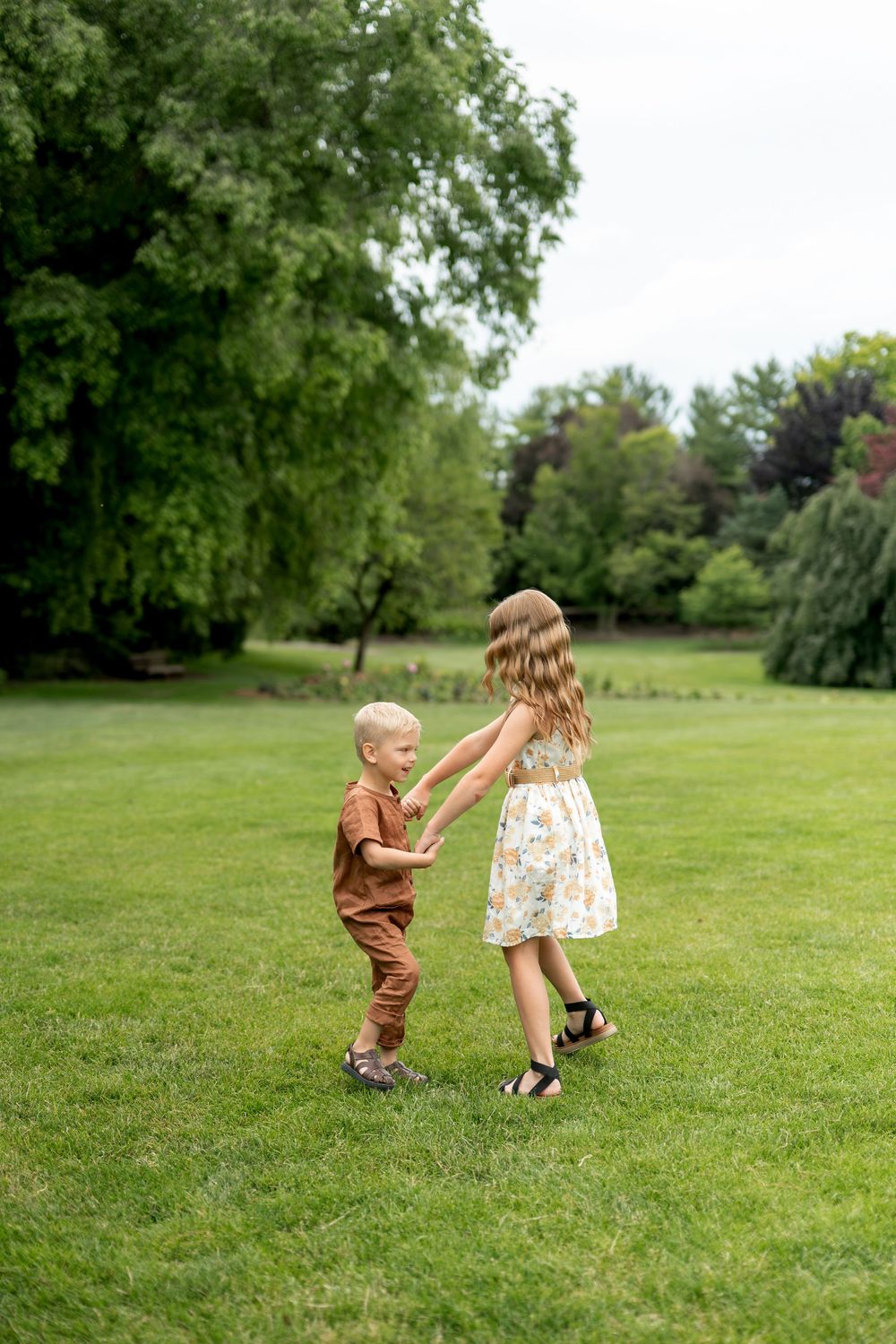 Two young children hold hands and spin together on a green lawn in a park.
