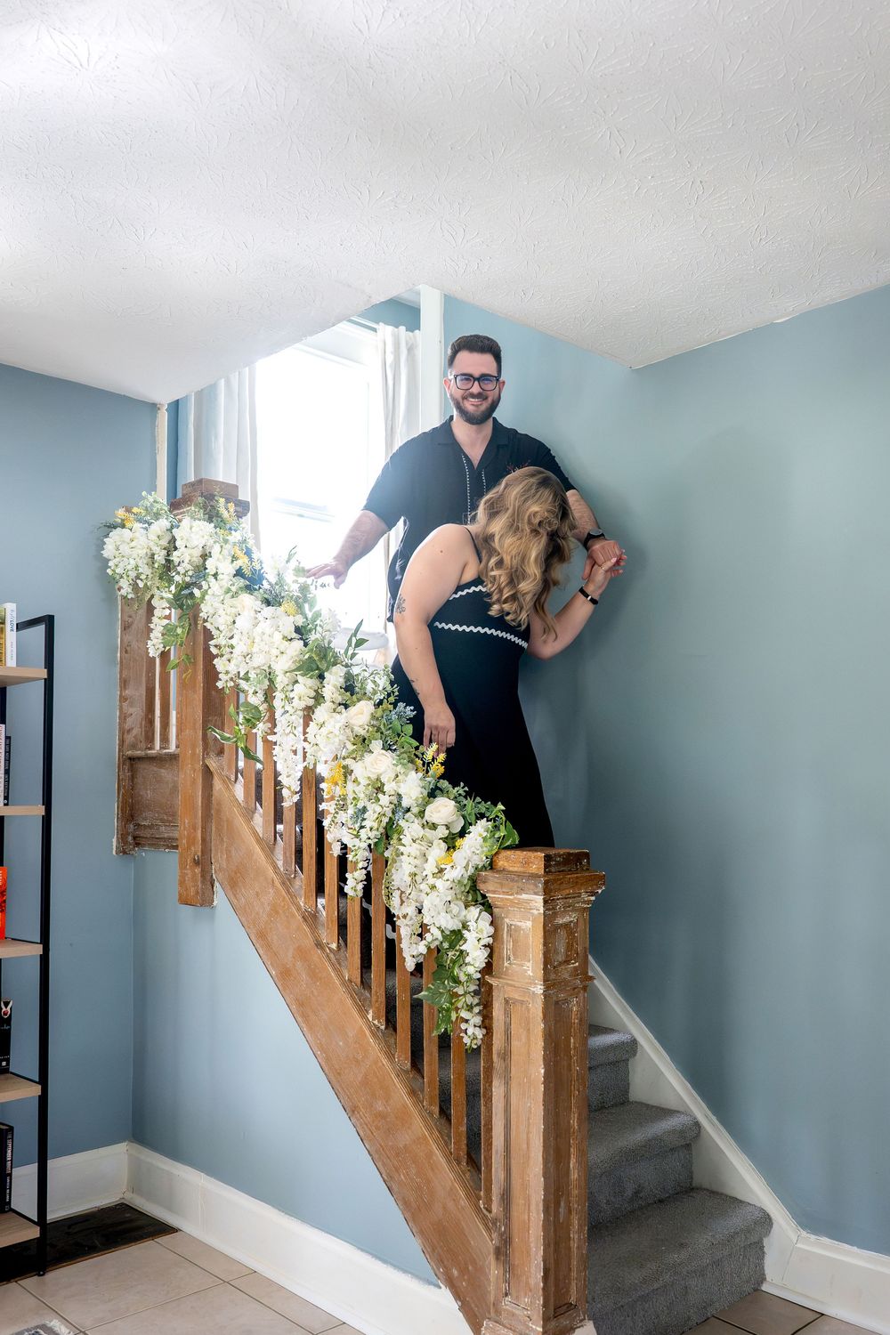 Couple in black outfits posing on floral-decorated wooden staircase with blue walls.