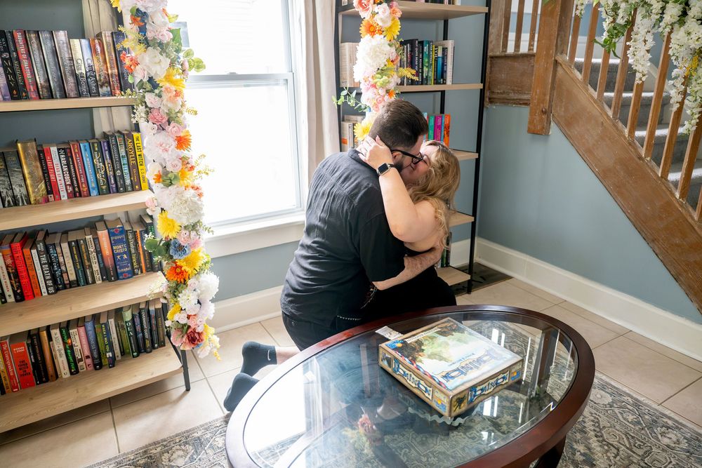 Couple embracing after a proposal in a sunlit room with flower garlands and bookshelves filled with colorful books.