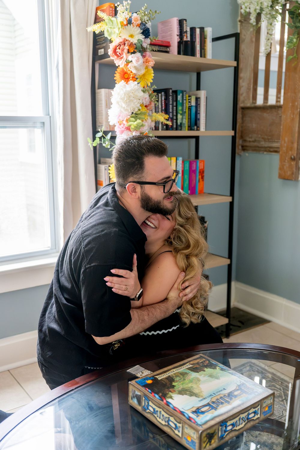 A couple shares a joyful embrace near a bookshelf with colorful floral decorations by a bright window.