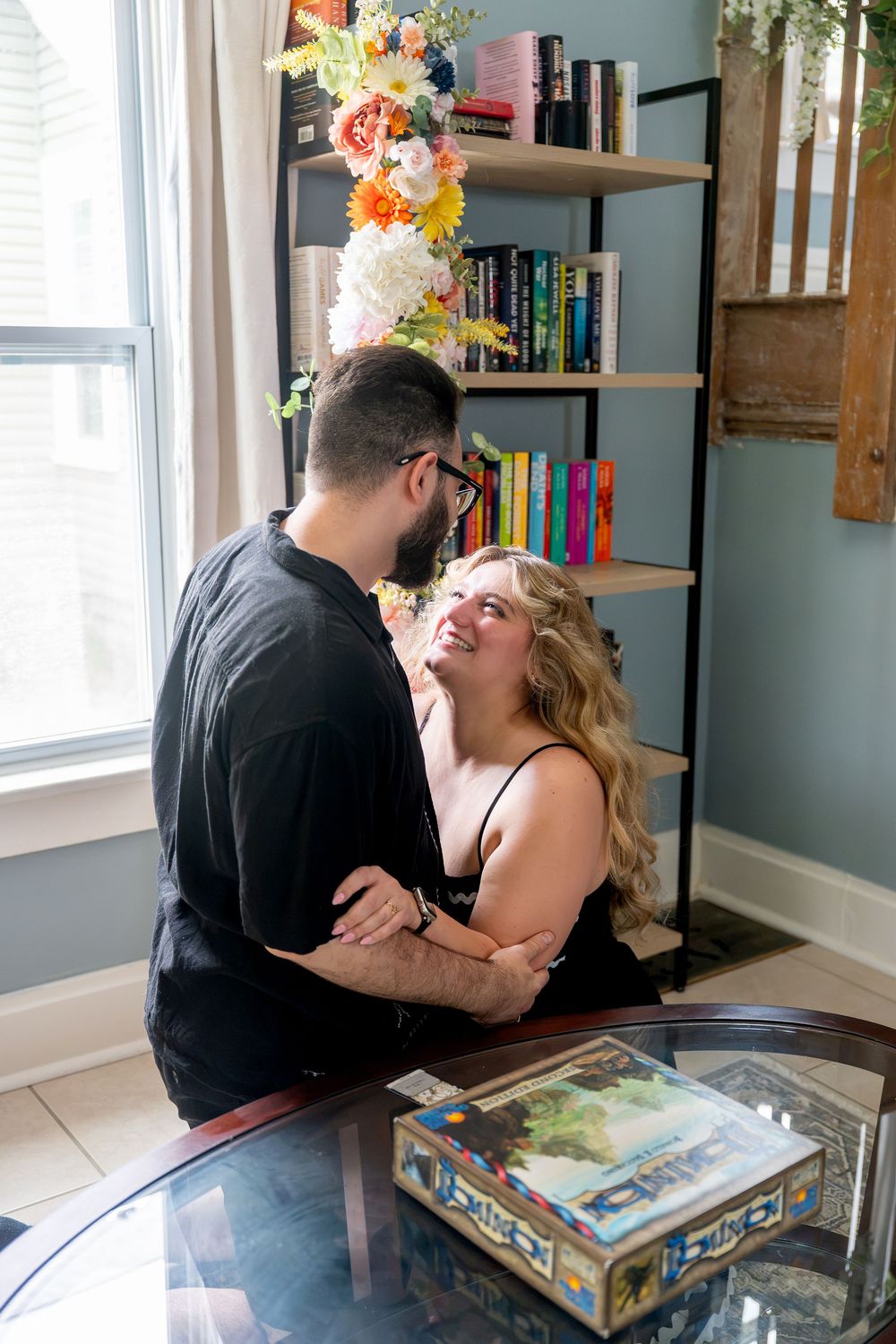 A smiling couple shares a tender moment by a bookshelf decorated with colorful flowers near a sunlit window.