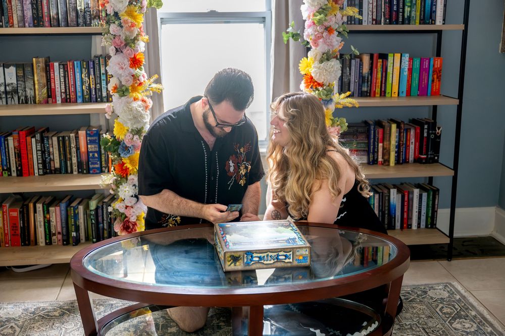 Couple playing board game together at glass coffee table in cozy home library with floral decorations.
