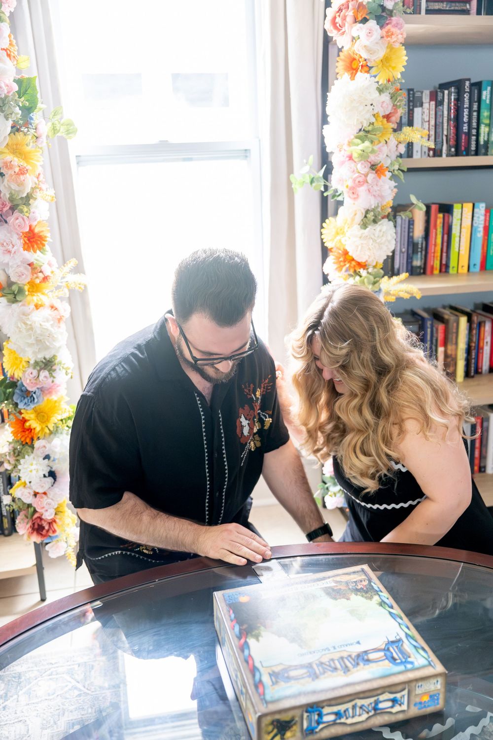 A couple leans over a glass coffee table examining a board game near a flower-adorned window with bookshelves behind them.