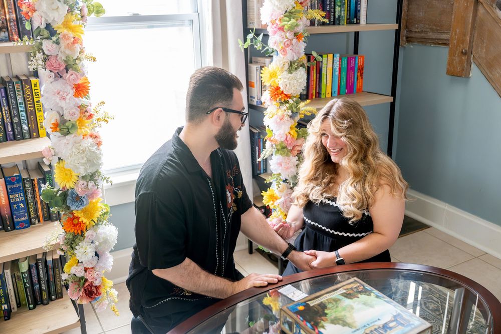 Man and woman sharing a joyful moment at a glass table in a cozy room decorated with flowers and colorful bookshelves.