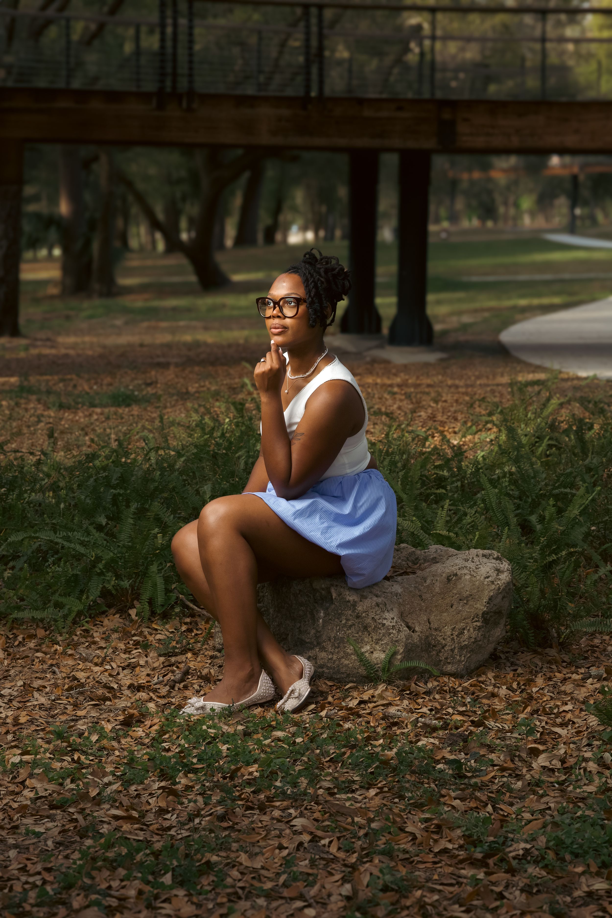 Young Black woman in glasses sits on a rock in a park, wearing a white top and blue skirt, looking thoughtful.