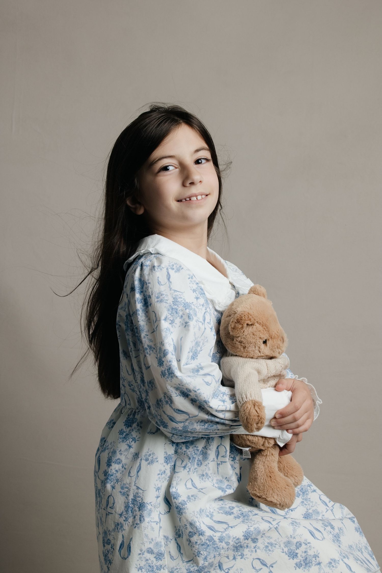 Young girl in floral blue dress hugging a teddy bear, smiling warmly against a neutral background.