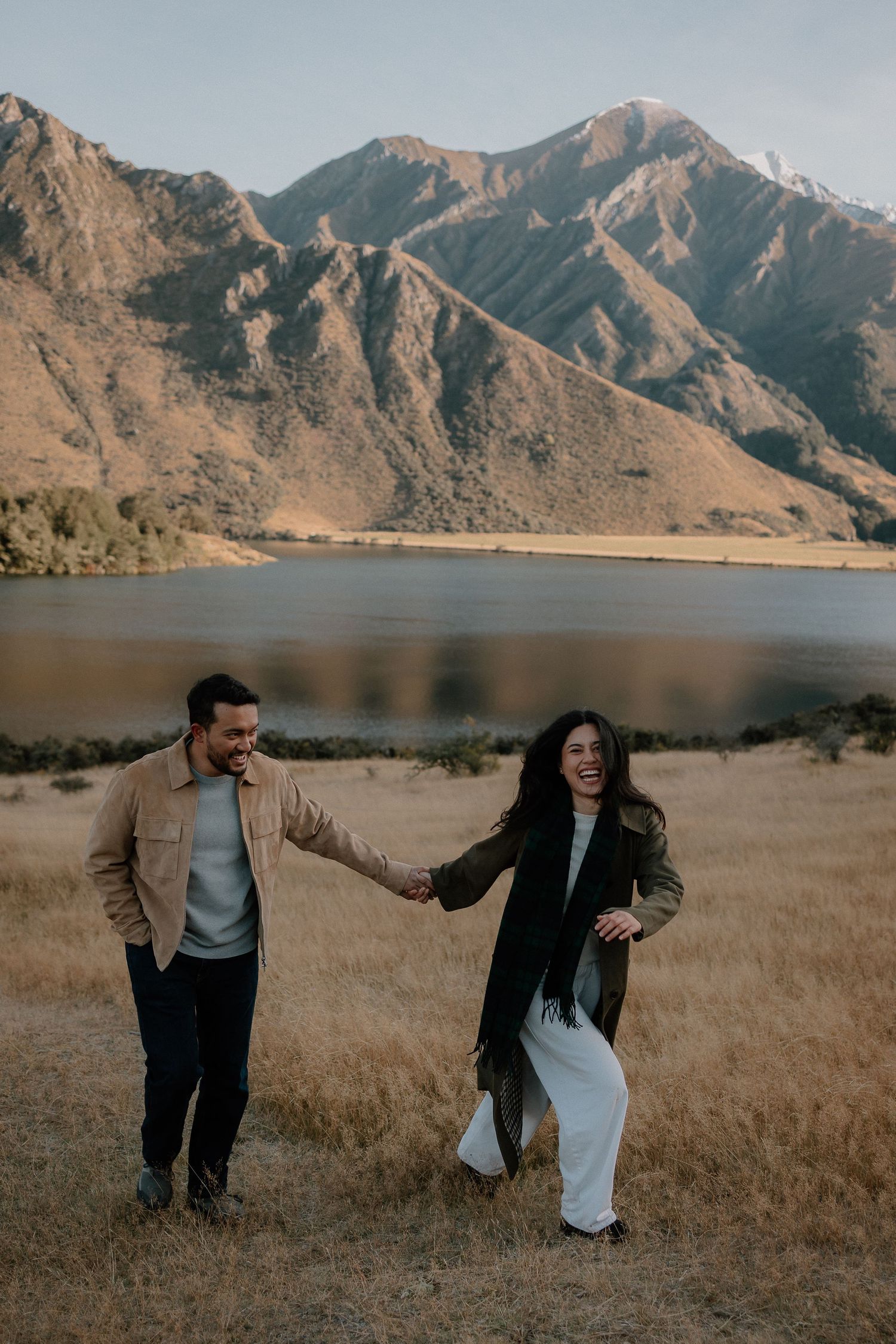 Couple holding hands laughing in a field with a mountain lake backdrop in autumn.