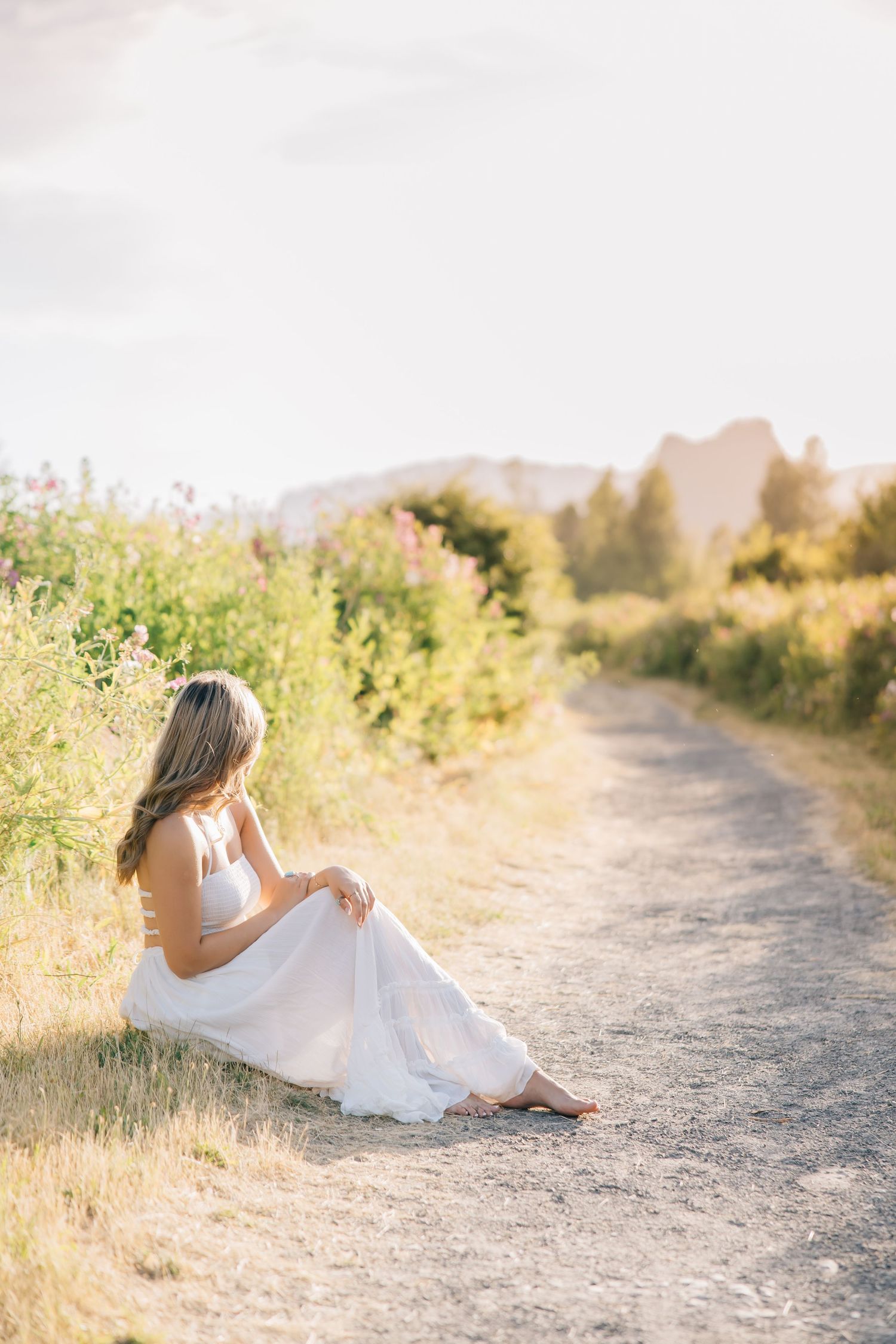A barefoot woman in a white dress sits peacefully on a rural path surrounded by wildflowers and golden sunlight.