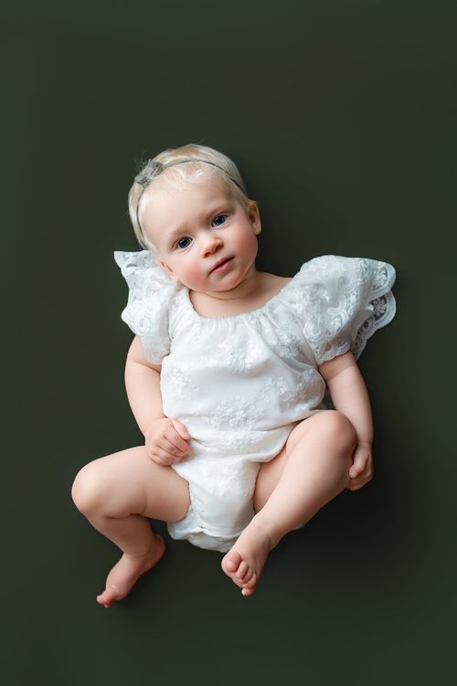 Baby girl in white lace romper lying on green backdrop, wearing headband, looking up at camera.