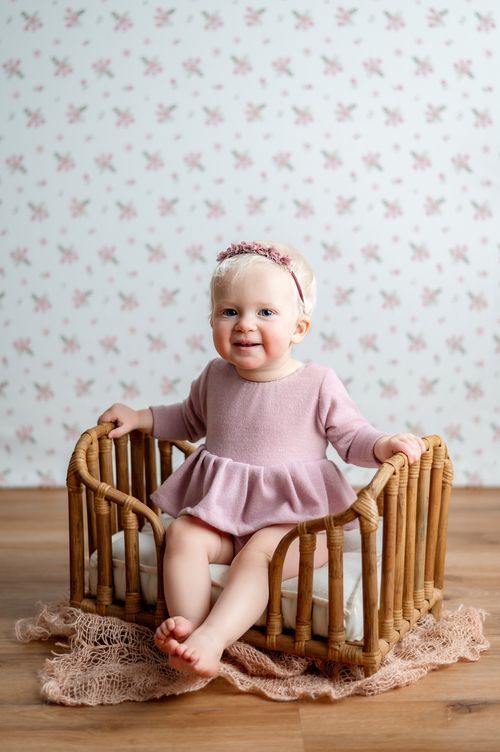 Smiling baby girl in pink dress sitting in a wicker crib with floral wallpaper background.