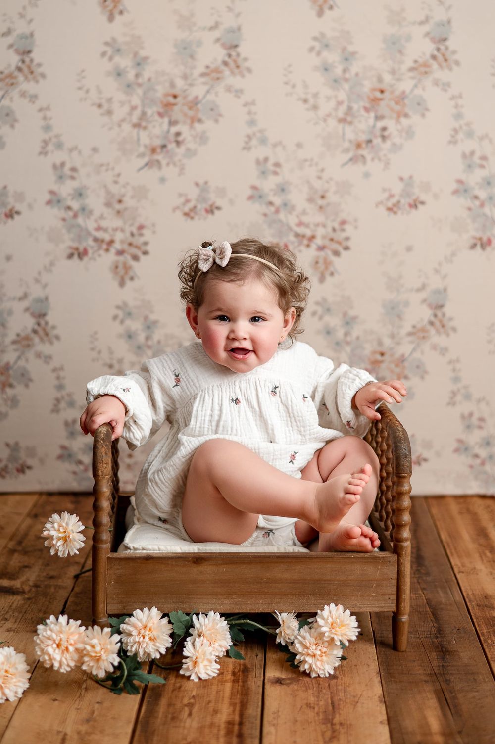 Smiling baby girl in white romper sits in a small wooden bed with flowers, against a floral wallpaper backdrop.