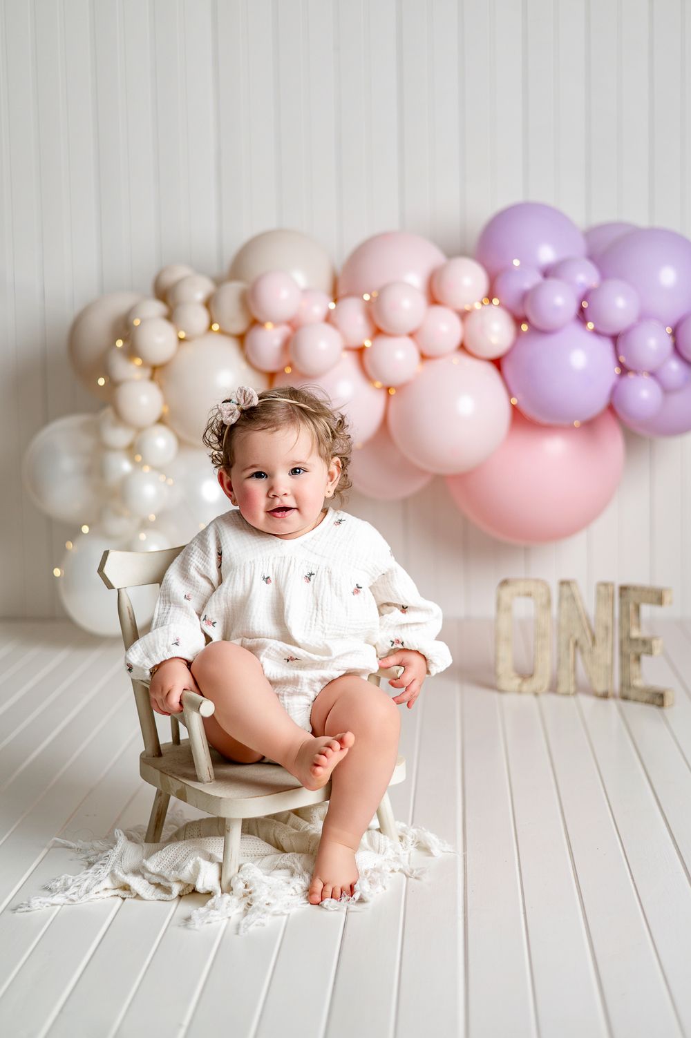 Smiling baby girl in white outfit sits on small chair surrounded by pink and purple balloon garland with ONE sign.