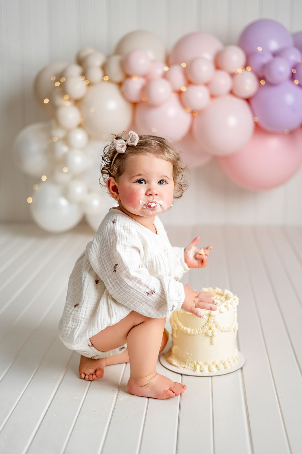 Baby girl in white dress smashes first birthday cake surrounded by pastel balloon garland backdrop.