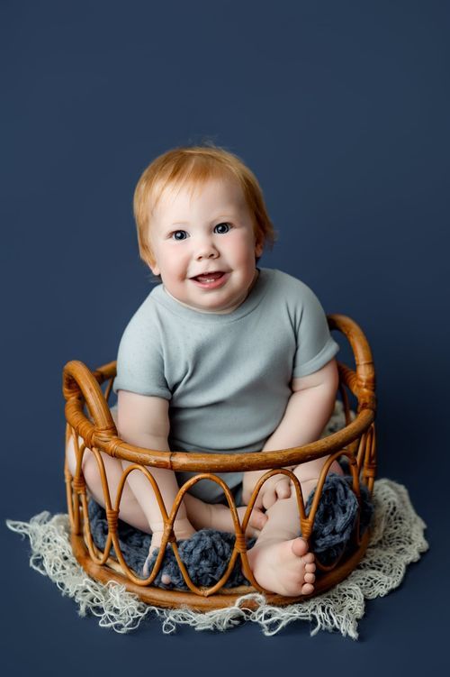 Smiling red-haired baby sitting in a wicker basket with blue blanket on a navy background.