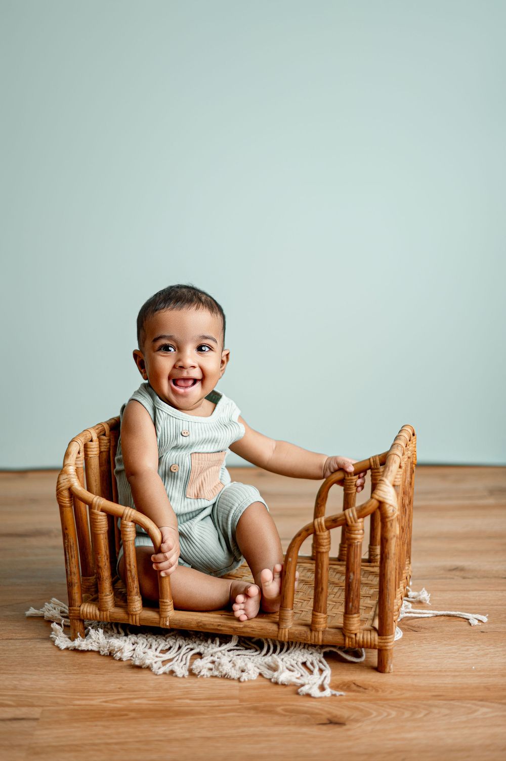 Smiling baby in green romper sitting in a small rattan crib on a wood floor against a mint green background.