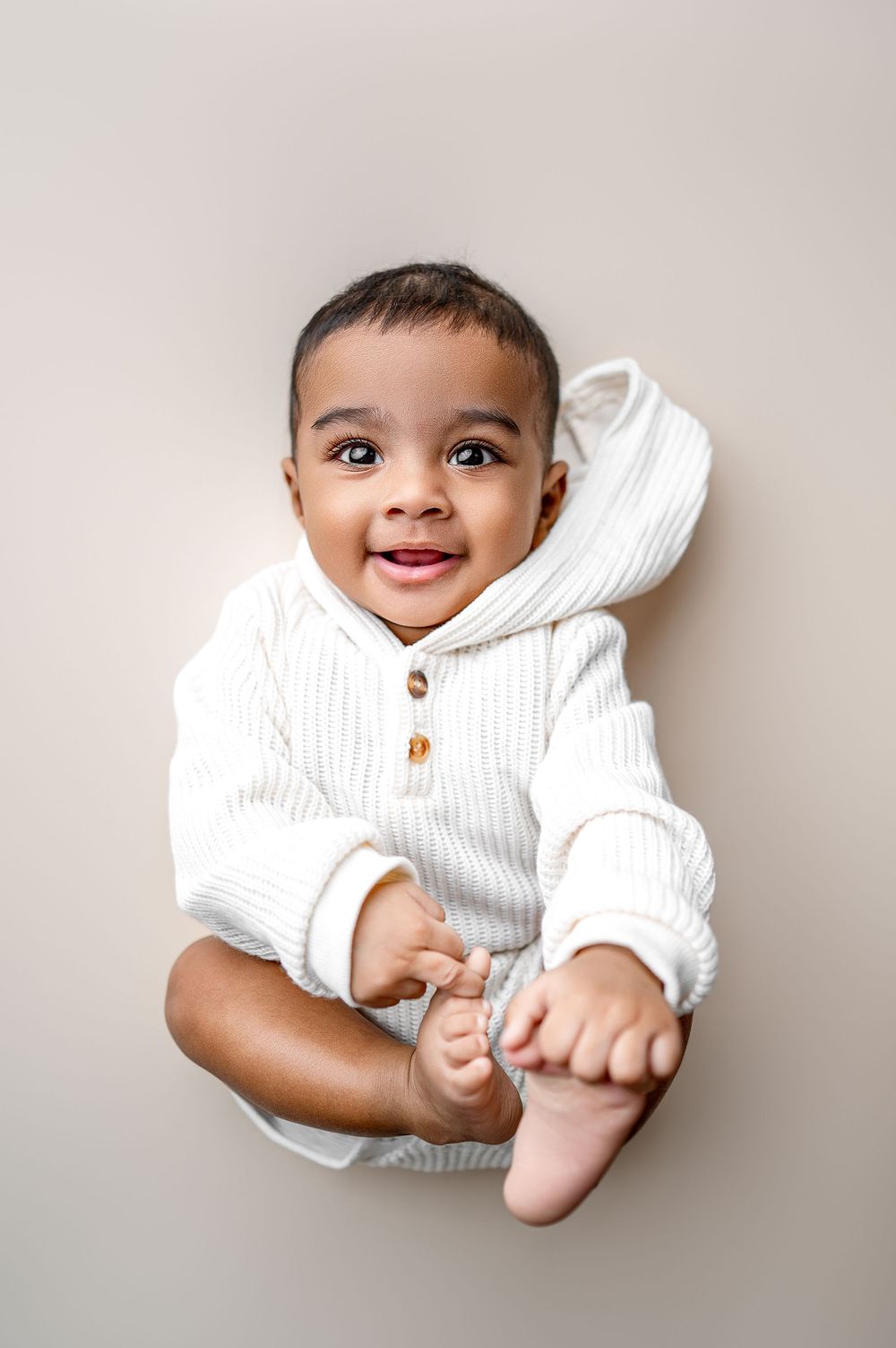 Happy African American baby in white hoodie romper lying down holding feet, smiling at camera.