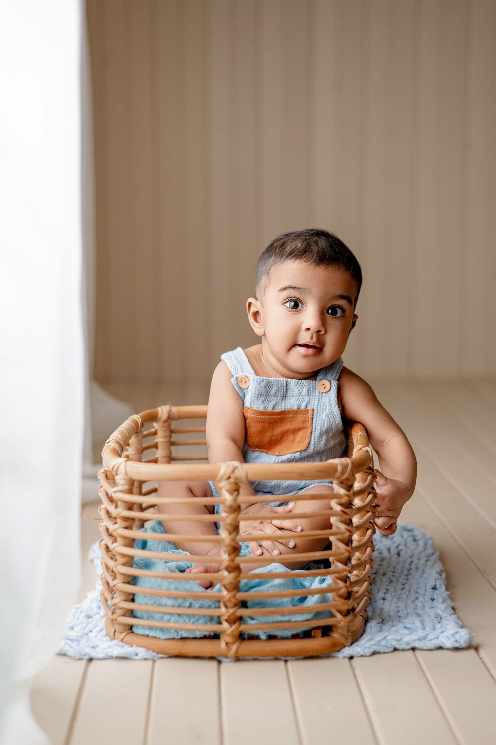 Adorable baby sitting inside a wicker basket on a light blue blanket, wearing a gray linen romper, looking at camera.