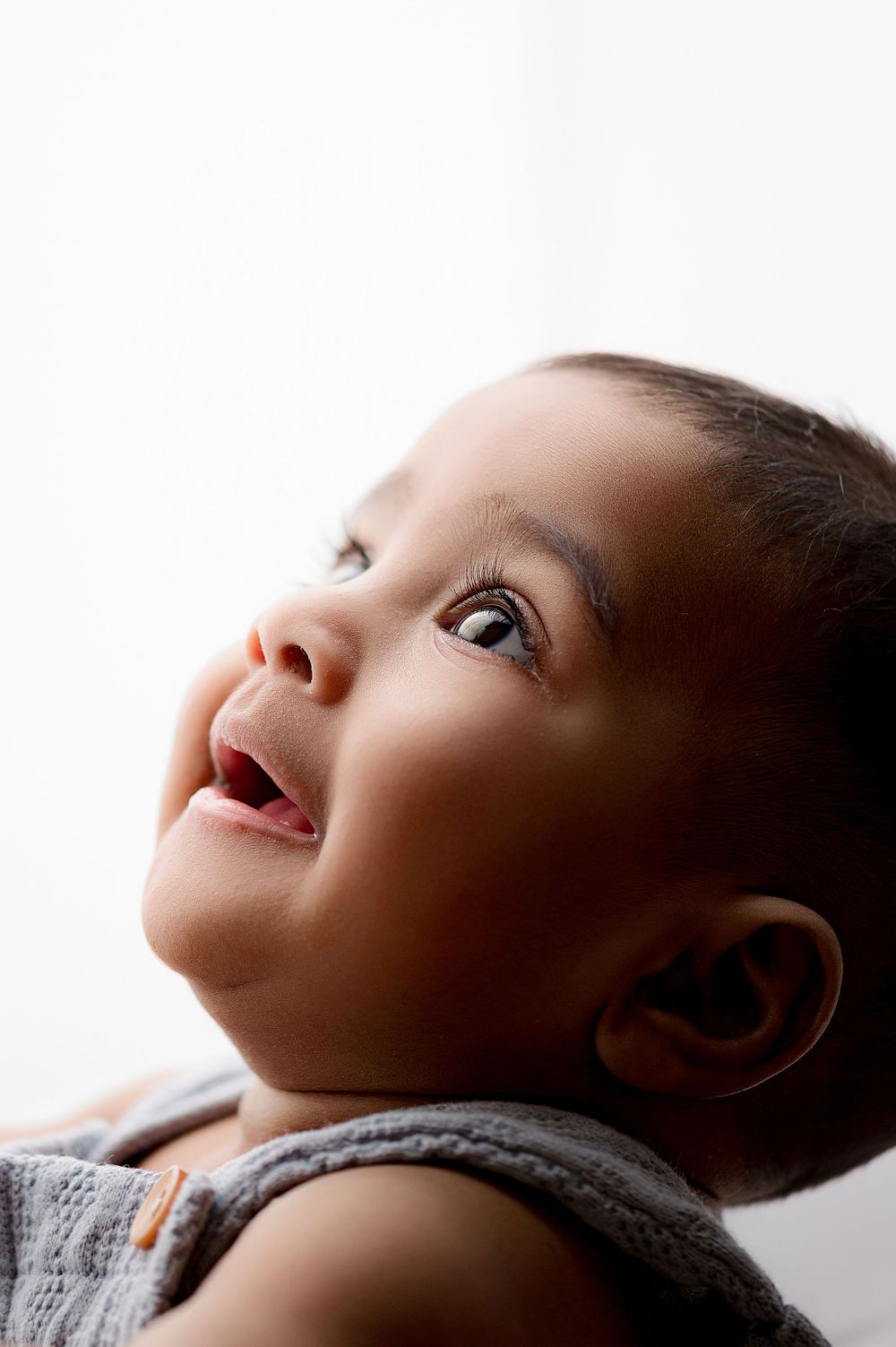 A curious baby with dark eyes looks upward with a slight smile, wearing a gray sleeveless outfit against a white background.