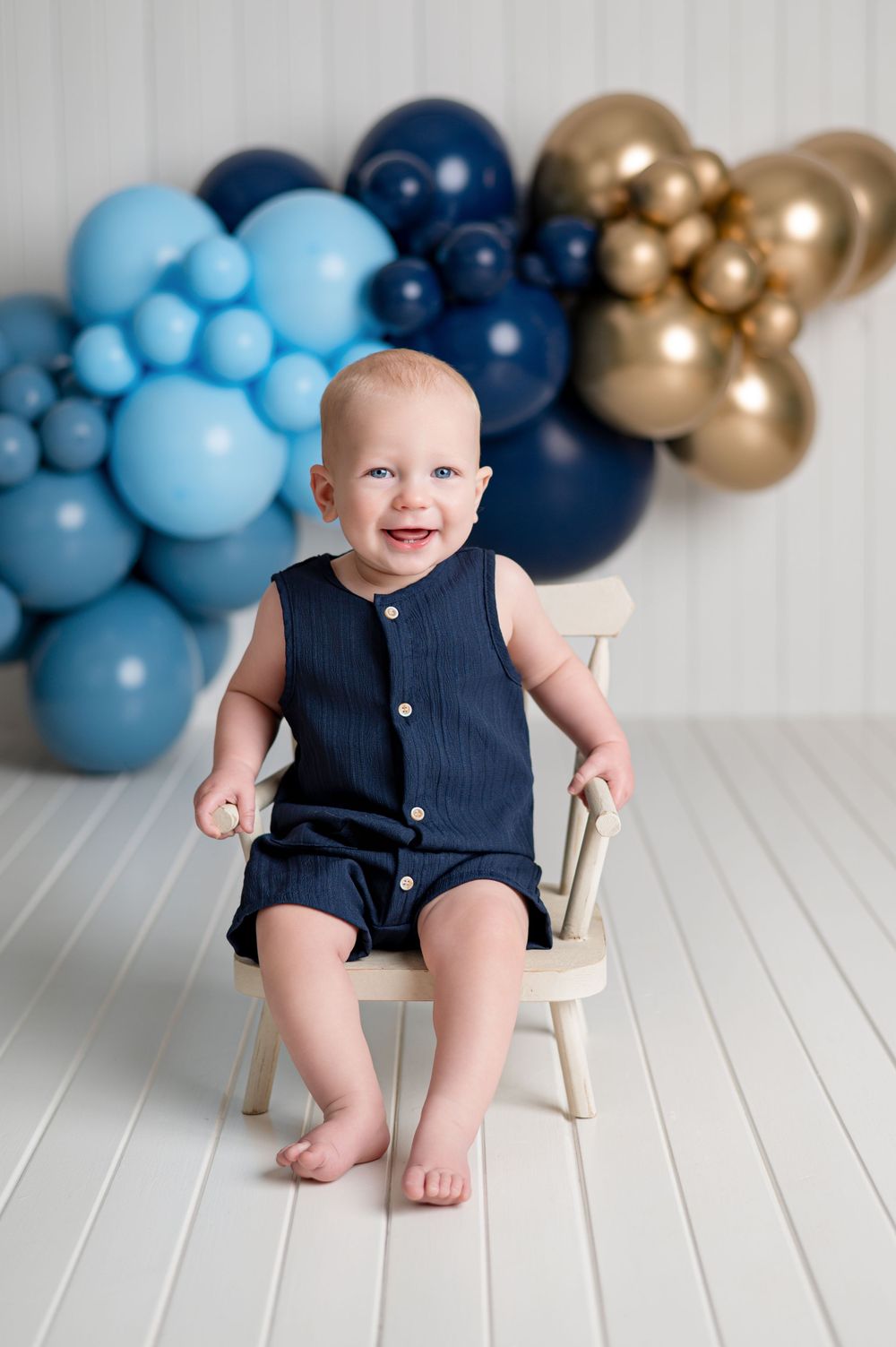 A smiling baby boy in a navy romper sits on a small wooden chair with blue and gold balloons in the background.