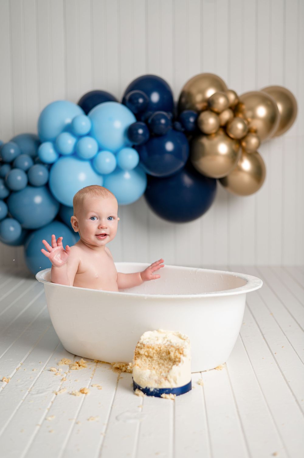 Baby in white tub at cake smash party with blue and gold balloon garland backdrop.