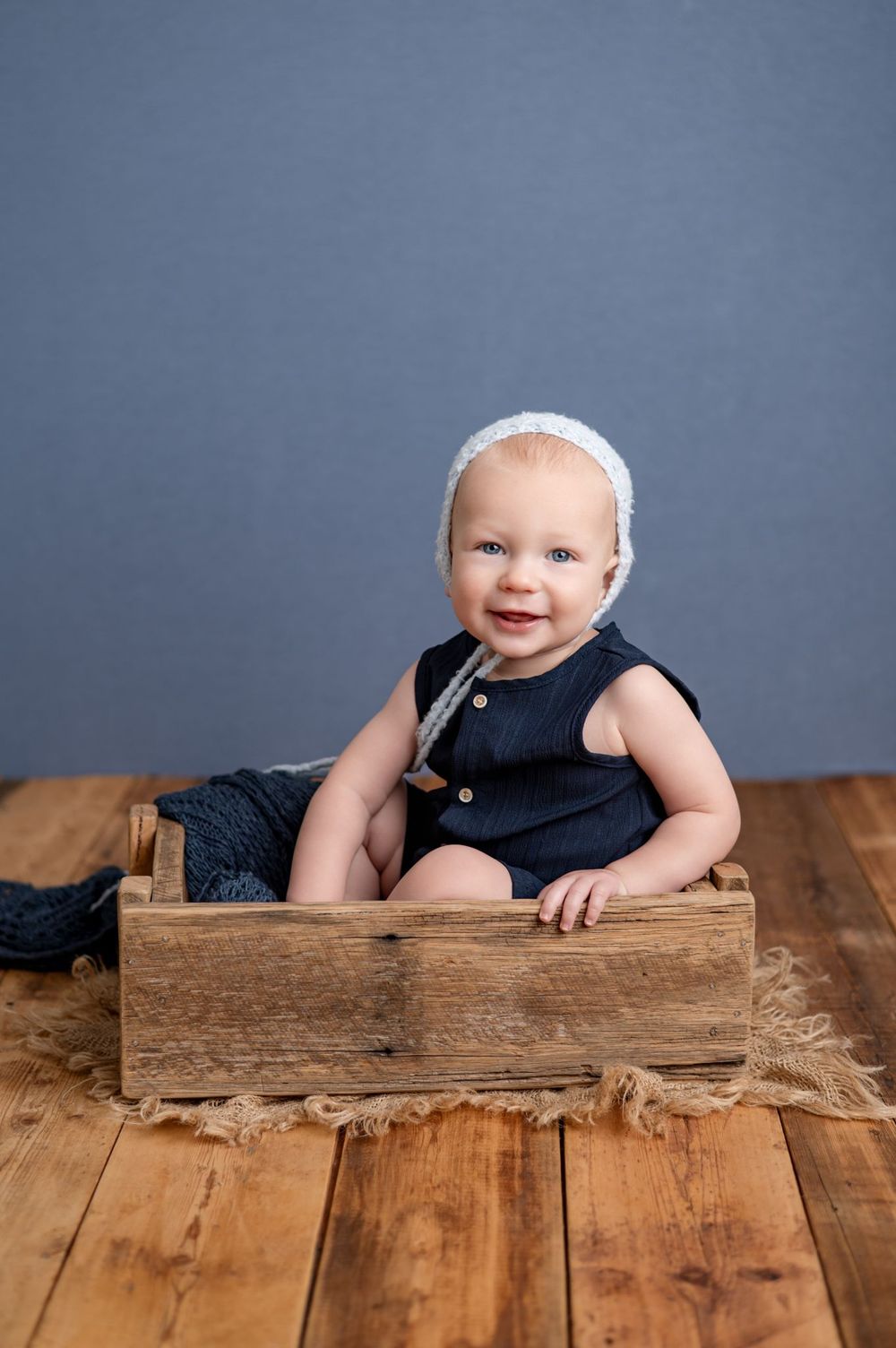 Smiling baby in navy romper and white bonnet sitting in rustic wooden crate on hardwood floor.