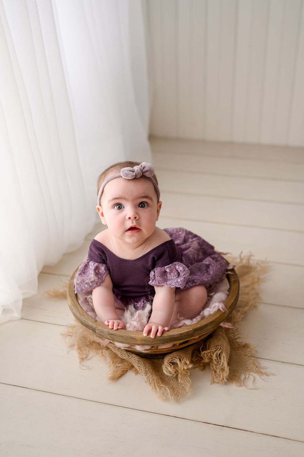 Baby girl in purple romper and bow headband sits in wooden bowl prop for professional studio portrait photo.