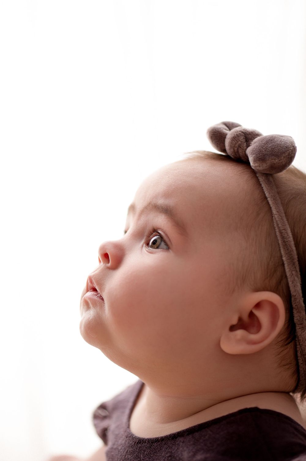 Adorable baby girl wearing a brown bow headband looks up curiously with big eyes and chubby cheeks.