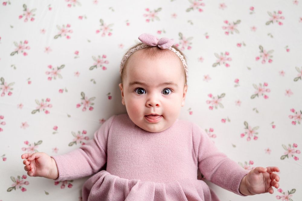 Adorable baby girl in pink dress with floral headband lying on floral background, looking up with bright blue eyes.