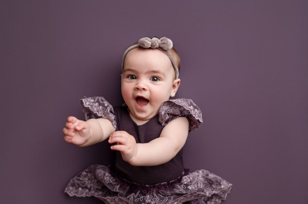 Adorable baby girl in purple ruffled dress and bow headband laughing joyfully during professional studio portrait session.