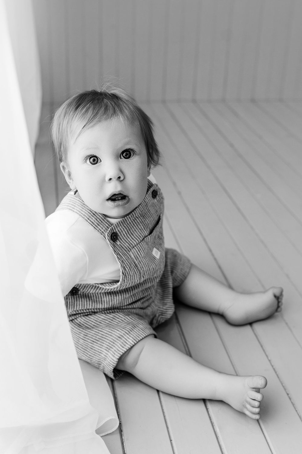 Black and white photo of a curious baby sitting on a wooden floor wearing a striped romper.