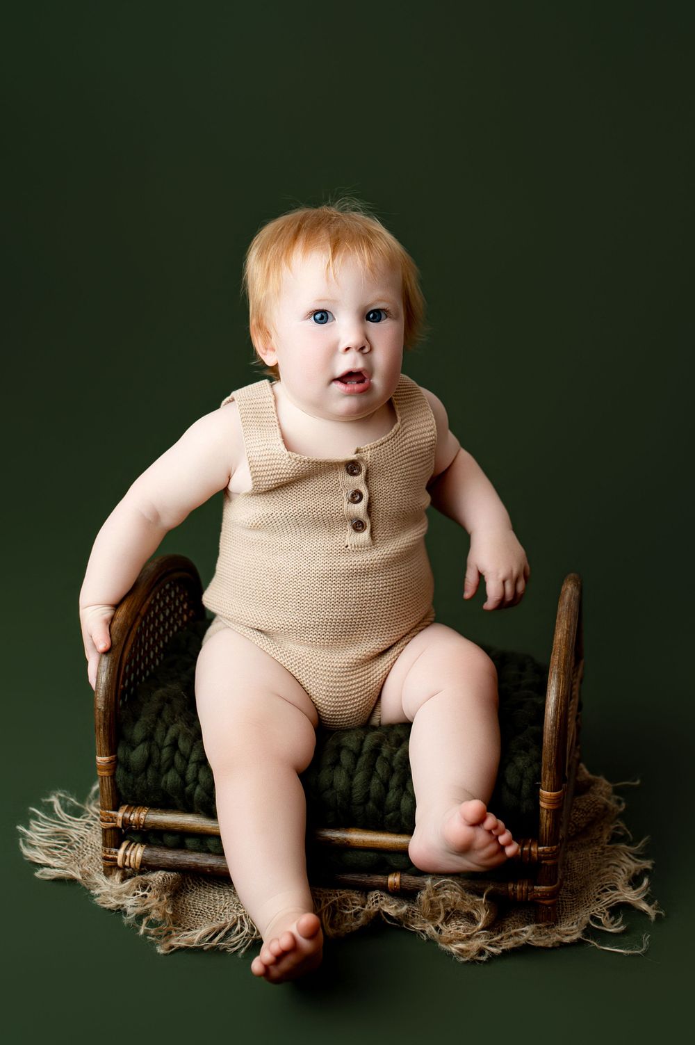 Adorable red-haired baby in beige knit romper sitting on small wooden chair against green background.