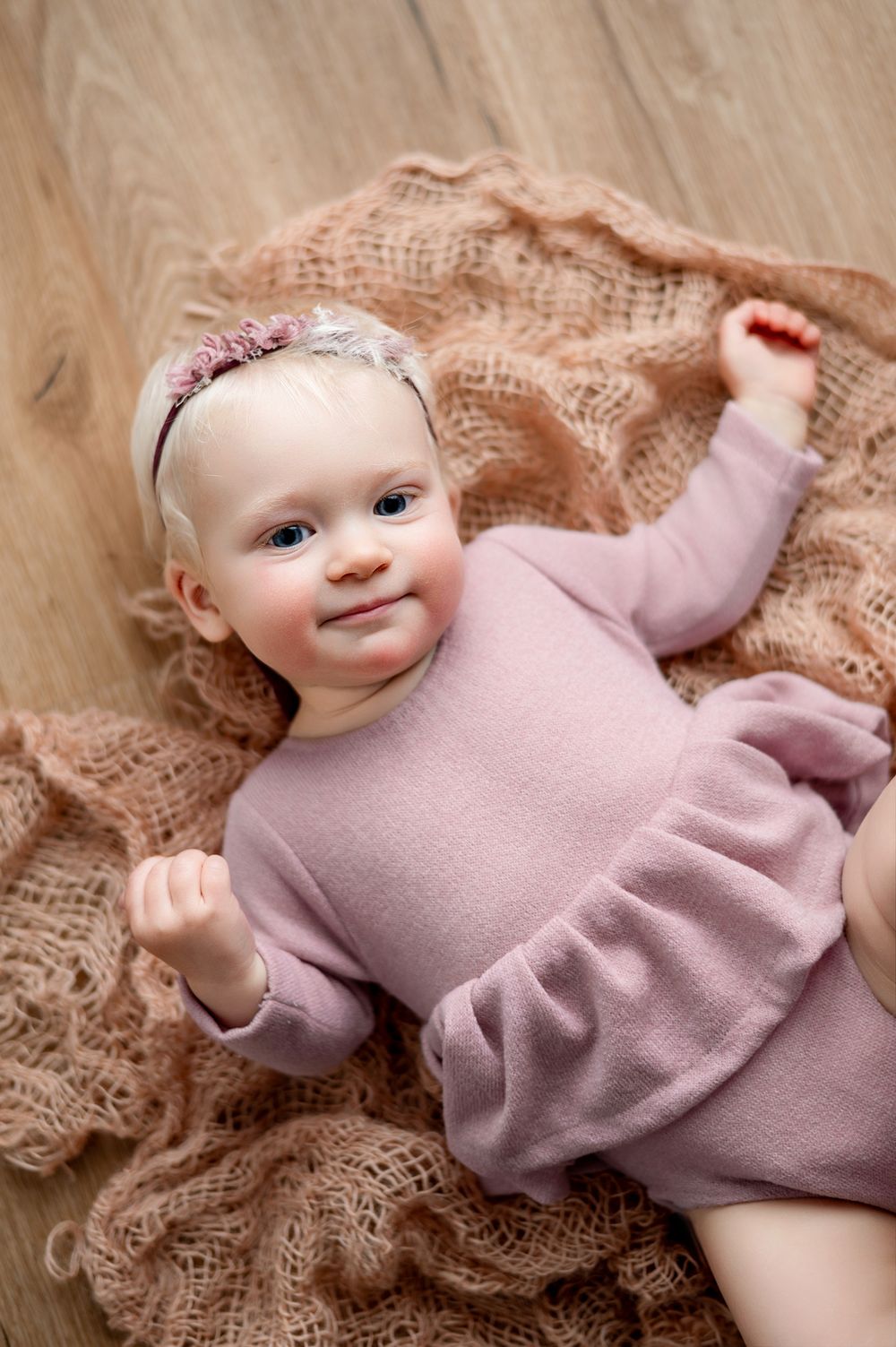 A smiling baby girl in a pink ruffle romper and floral headband lying on a beige knit blanket on a wood floor.