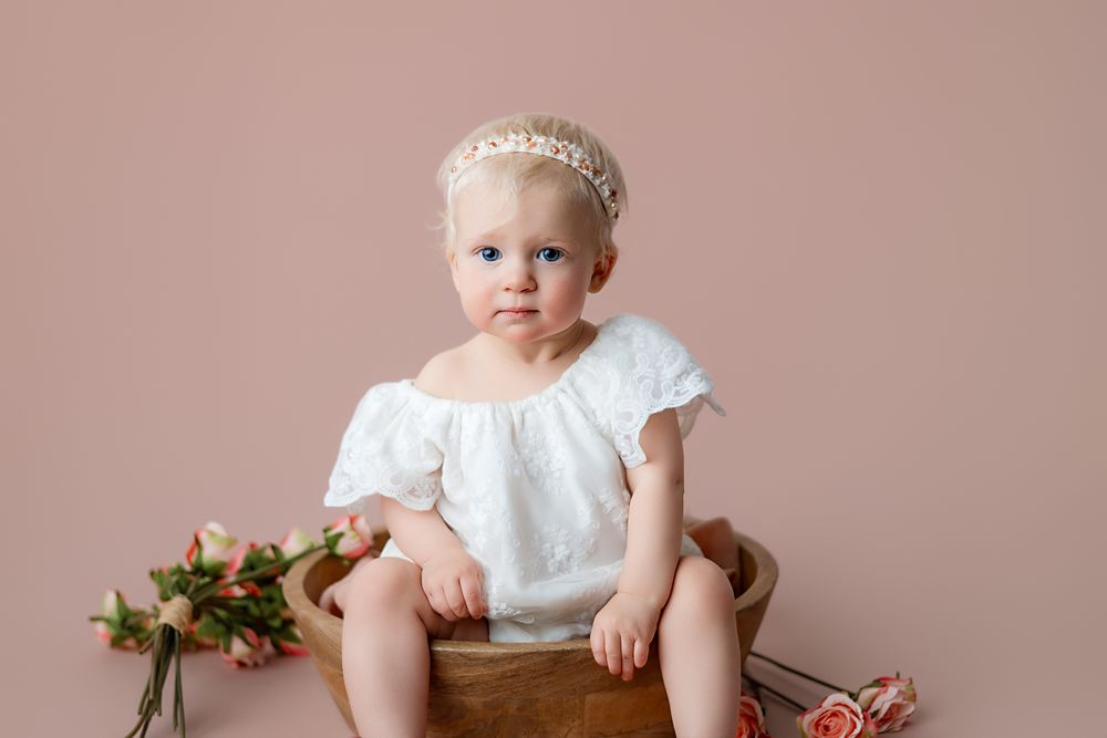 A baby girl in a white dress and floral headband sits in a wooden bowl surrounded by pink roses on a blush background.